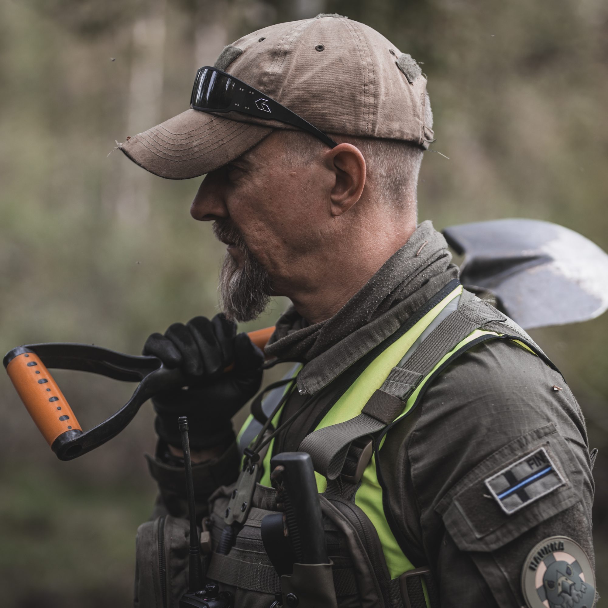 Lähi kuva miehestä joka katsoo sivulle. Päässään ruskea lippalakki ja aurinkolasit. Lapioa hän kannattelee olallansa.