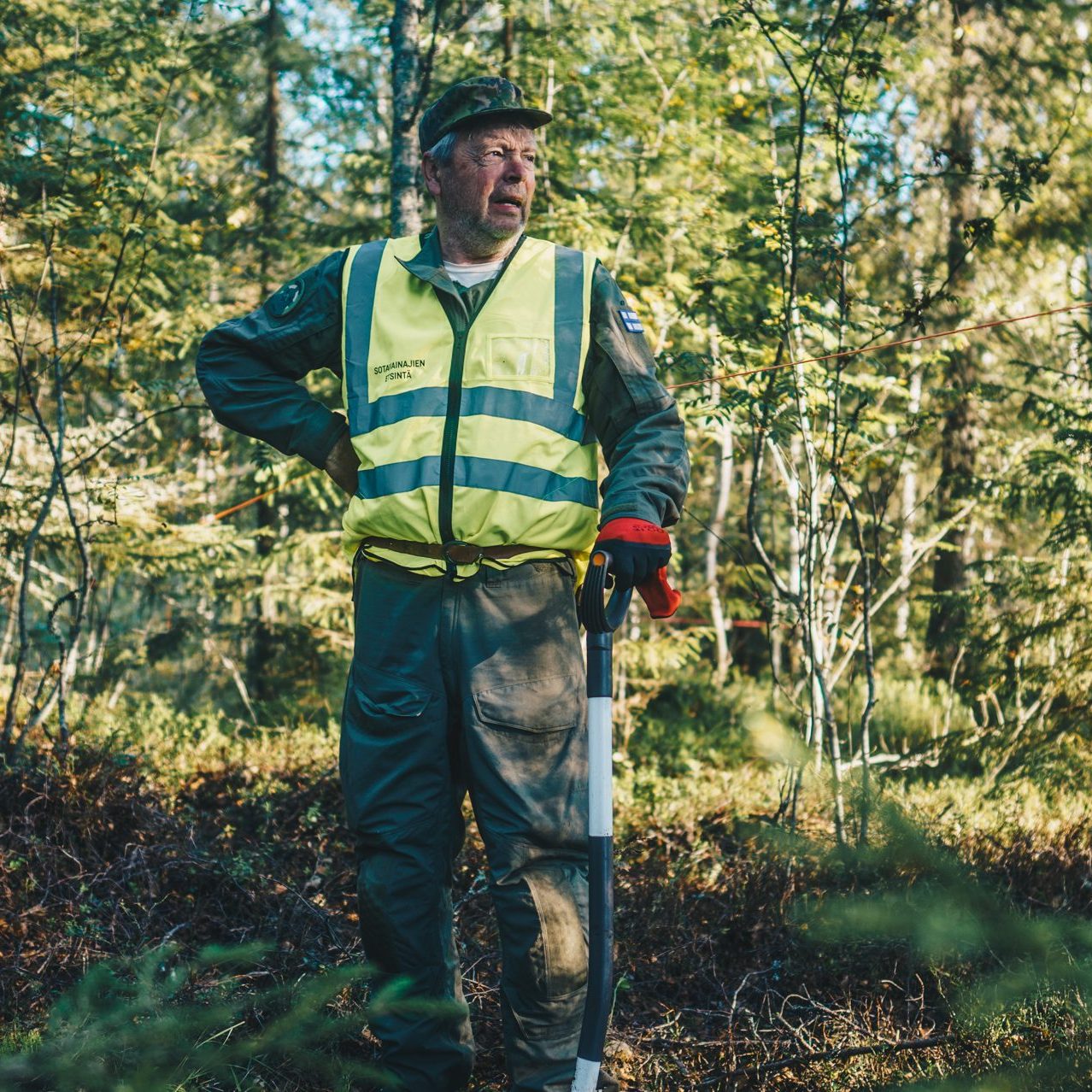 Mies seisoo metsässä huomioliiveihin pukeutuneena jossa lukee "Sotavainajien etsintäryhmä". Hänellä on käsissään lapio.