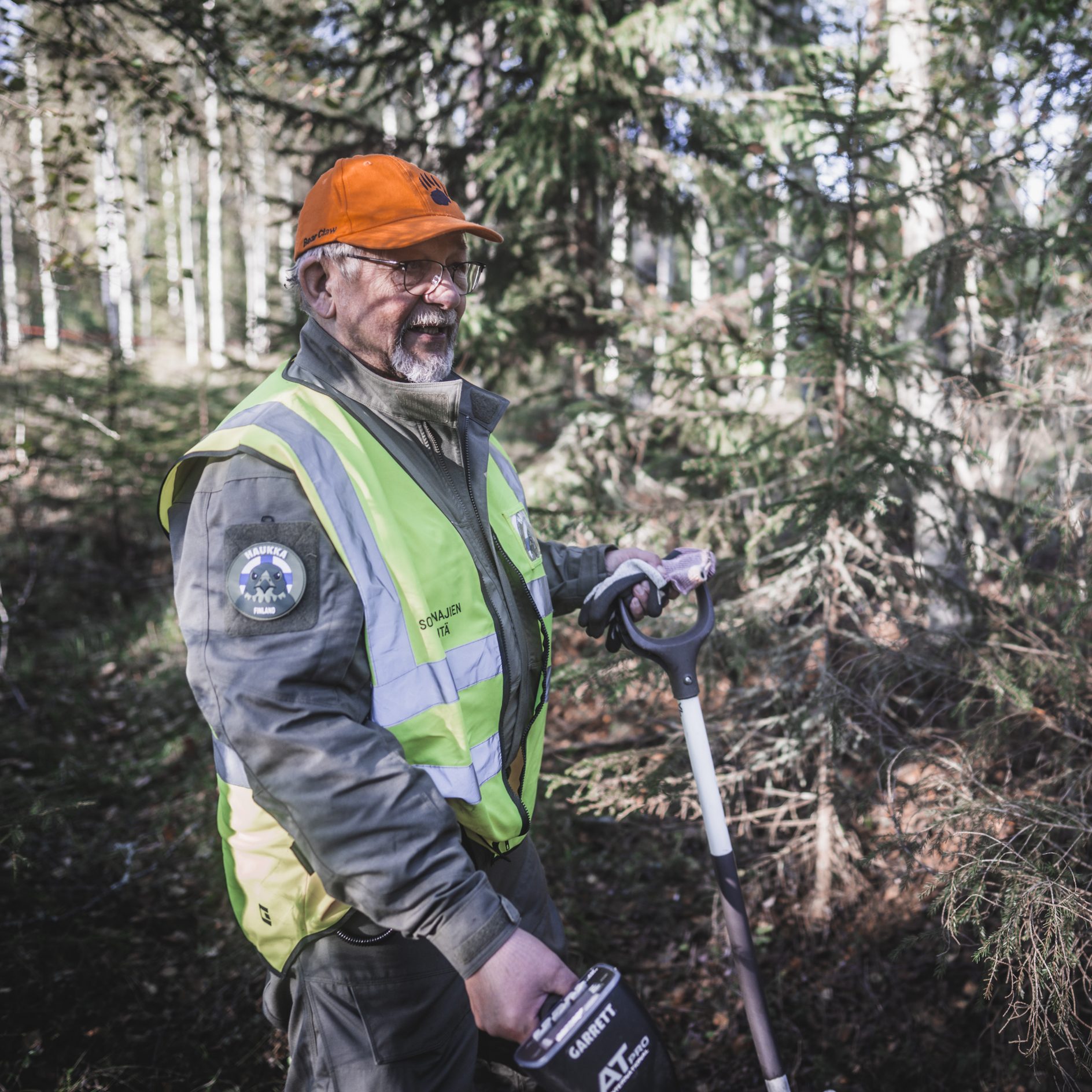 Mies seisoo metsässä huomioliiveihin pukeutuneena jossa lukee "Sotavainajien etsintäryhmä". Hänellä on käsissään metallinpaljastin ja lapio.