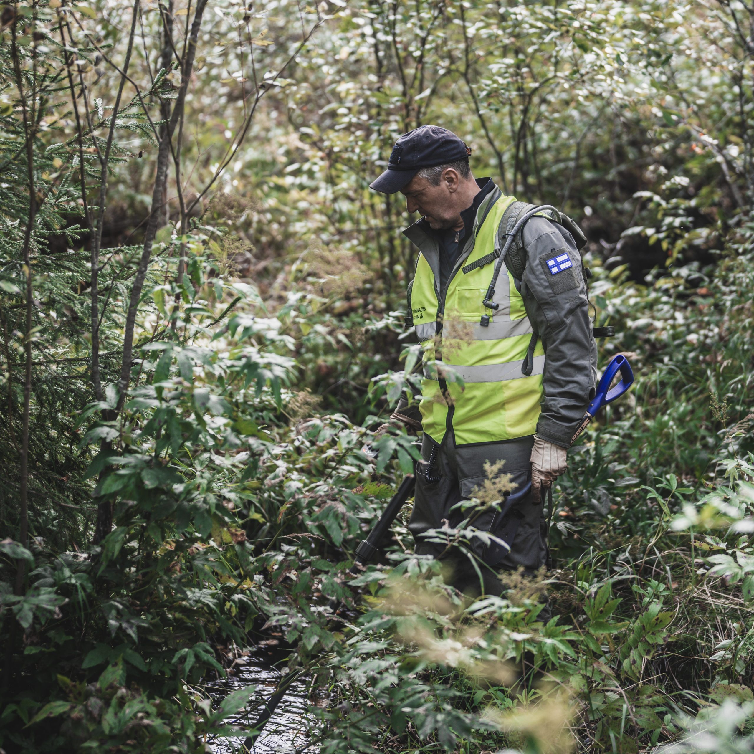 Mies tekee etsintää metsässä huomioliiveihin pukeutuneena jossa lukee "Sotavainajien etsintäryhmä". Hänellä on käsissään metallinpaljastin ja lapio.