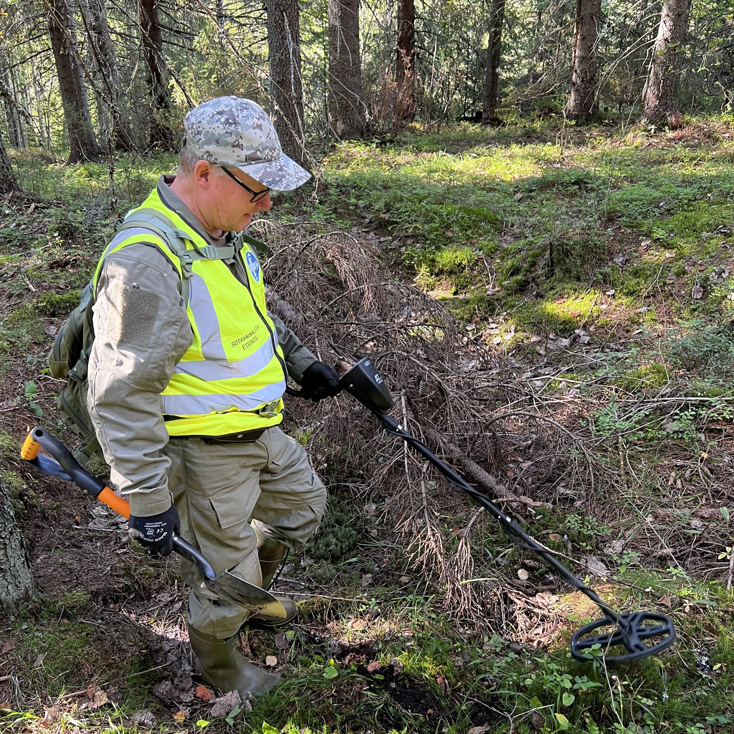 Mies tekee etsintää metsässä huomioliiveihin pukeutuneena jossa lukee "Sotavainajien etsintäryhmä". Hänellä on käsissään metallinpaljastin ja lapio.