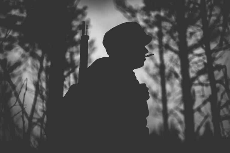 Sotilaalla on suussa tupakka ja selässään kivääri. Siluetti jossa on mustaa ja valkoista. Taustala on metsä.