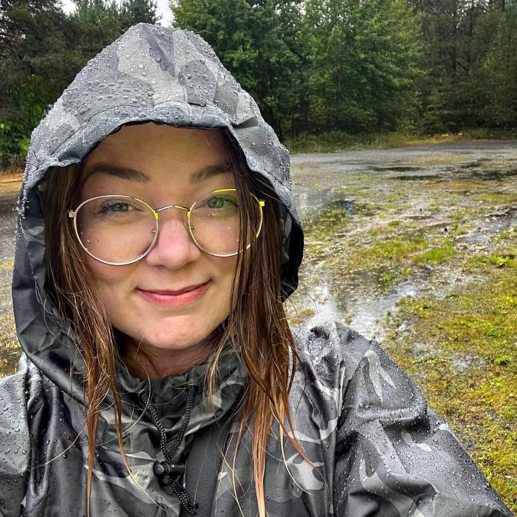 Nainen jolla on kultaisilla kehyksillä varustetut silmälasit seisoo maastokuvioinen takki päällä sateessa. Taustalla on metsää.