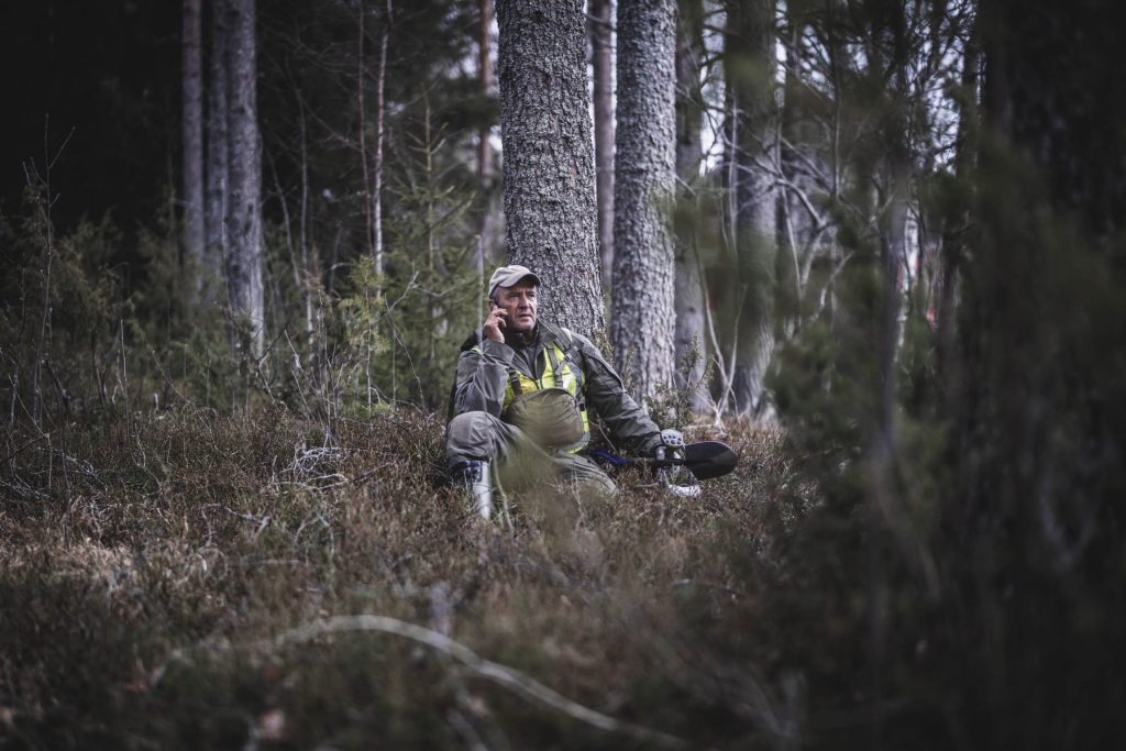 Etsijä istuu metsässä mättäällä nojaten puuhun ja puhuu puhelimessa.
